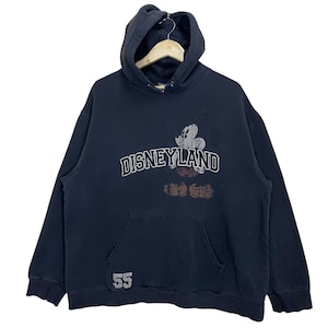 Puede incluir: Sudadera con capucha negra con un gr&aacute;fico de Mickey Mouse de estilo vintage y el texto "DISNEYLAND" en blanco. La sudadera tiene un gran bolsillo delantero y el n&uacute;mero "55" impreso en la parte inferior izquierda.