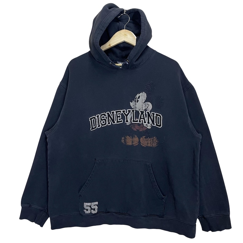 Puede incluir: Sudadera con capucha negra con un gr&aacute;fico de Mickey Mouse de estilo vintage y el texto "DISNEYLAND" en blanco. La sudadera tiene un gran bolsillo delantero y el n&uacute;mero "55" impreso en la parte inferior izquierda.