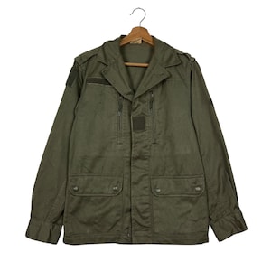 May include: Olive green military-style jacket with a collared neckline, two chest pockets, and two lower pockets. The jacket has a button-up front, epaulets, and patches on the sleeves. It is hanging on a wooden hanger.