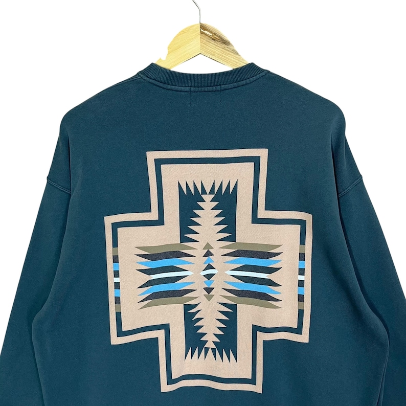 以下が含まれることがあります： 背中に大きなベージュの十字デザインが施された、ティール色のスウェットシャツ。 十字は、青、茶色、白のアクセントが付いた幾何学模様が特徴です。 スウェットシャツはクルーネックと長袖です。