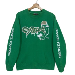 May include: Green sweatshirt with the words "DANCE TO MUSIC" printed vertically on the sleeves. The front features graffiti-style text and a cartoon figure holding a spray can. The sweatshirt is hanging on a wooden hanger.