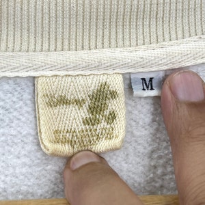 K&ouml;nnte beinhalten: Ein wei&szlig;es und gr&uuml;nes gewebtes Etikett mit dem Buchstaben "M" auf einem wei&szlig;en Etikett. Das Etikett ist an einem grauen Sweatshirt befestigt.