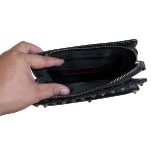 May include: Black leather clutch with gold-tone pyramid studs and a zippered closure. The interior is black with a red label. The clutch is open, revealing the inside pockets.