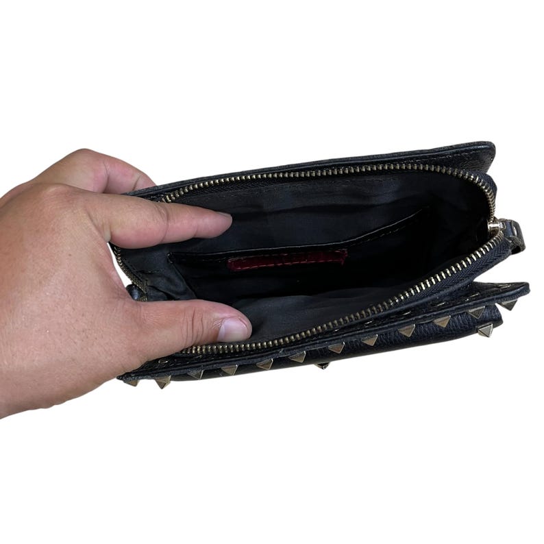 May include: Black leather clutch with gold-tone pyramid studs and a zippered closure. The interior is black with a red label. The clutch is open, revealing the inside pockets.
