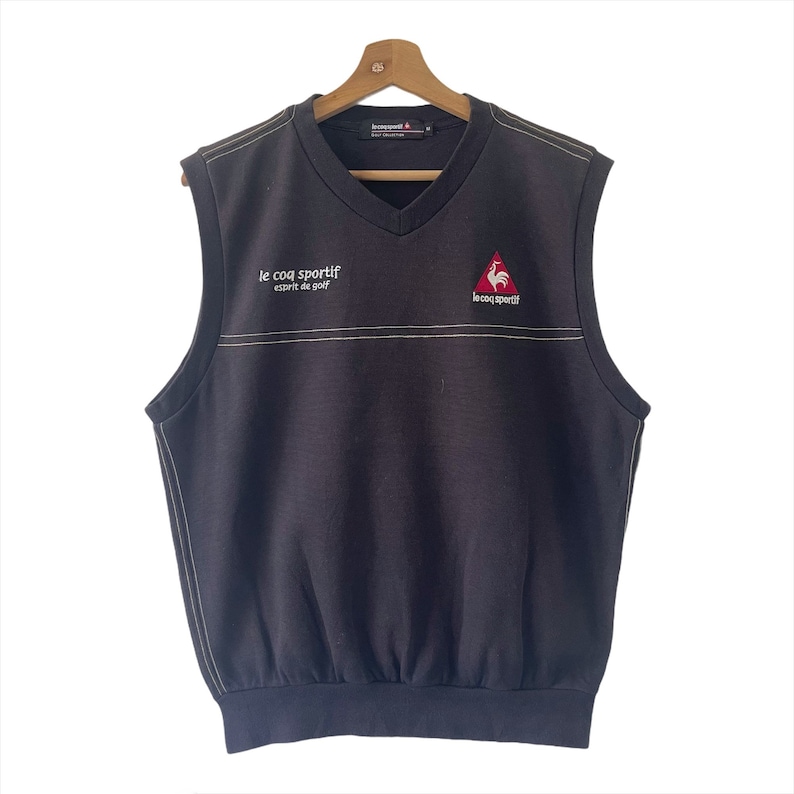May include: Black sleeveless sweater vest with a white embroidered logo that says "le coq sportif esprit de golf" and a red rooster logo. The vest has a V-neck and white stitching.