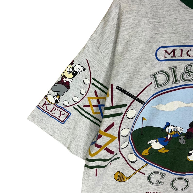 Puede incluir: Camiseta gris con Mickey Mouse y el Pato Donald jugando al golf. El dise&ntilde;o incluye las palabras "MICKEY DISNEY GOLF TOUR" y "KEY" en la manga. La camiseta est&aacute; hecha de un material suave y c&oacute;modo.