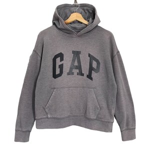 May include: A gray hoodie with the word "GAP" printed in large, black letters across the chest. The hoodie has a hood, a front pocket, and long sleeves. It is hanging on a wooden hanger against a white background.