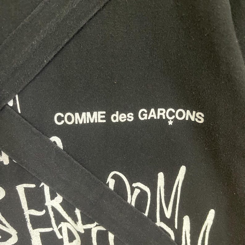 May include: Black t-shirt with white text that reads "Comme des Gar&ccedil;ons*" and "Freedom" in a graffiti style.