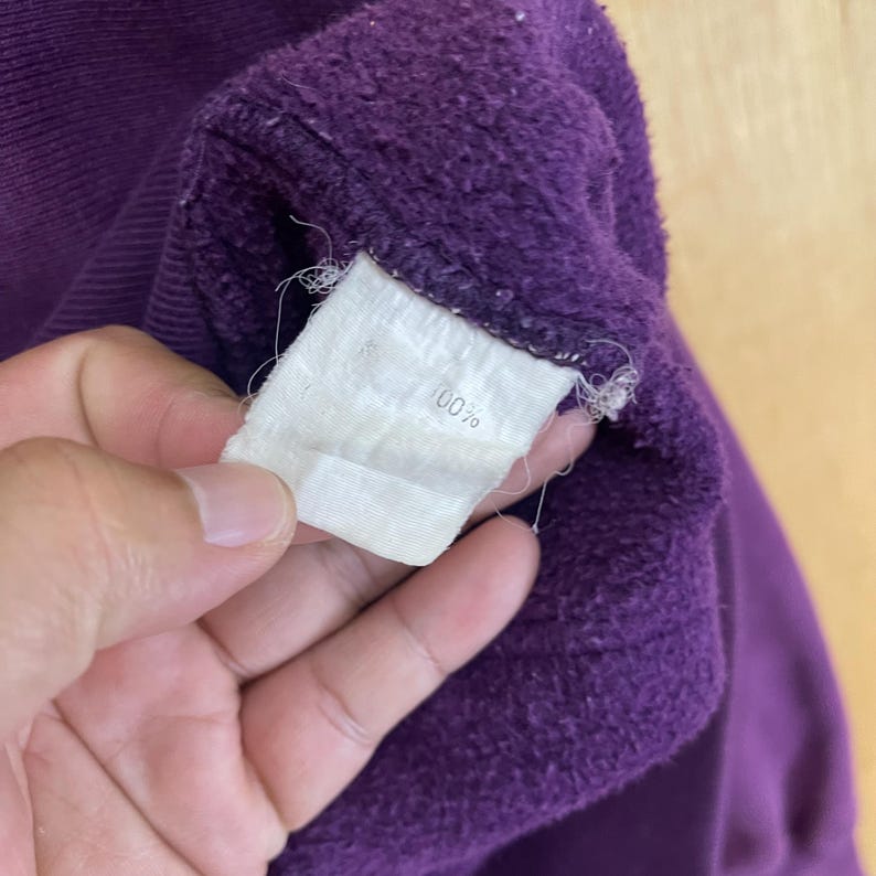 May include: Close-up of a deep purple sweatshirt. A white tag with the number "100%" is visible. The fabric appears soft and slightly worn.