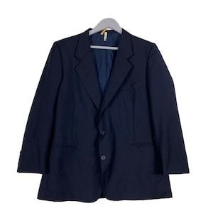 Puede incluir: Un blazer azul marino con cierre de dos botones, solapas con muescas y un bolsillo en el pecho. La chaqueta está colgada de una percha gris sobre un fondo blanco. El blazer tiene mangas largas y un diseño clásico y a medida.