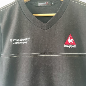 May include: Black v-neck t-shirt with a white embroidered logo that says "le coq sportif esprit de golf". The logo is on the left side of the shirt. On the right side of the shirt is a red embroidered logo of a rooster in a triangle with the text "le coq sportif" underneath.