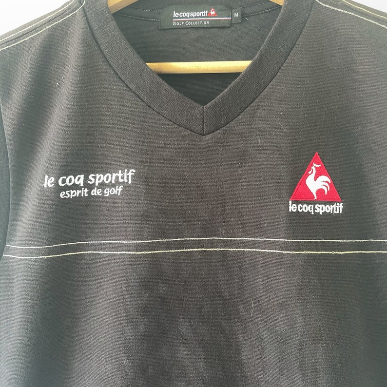May include: Black v-neck t-shirt with a white embroidered logo that says "le coq sportif esprit de golf". The logo is on the left side of the shirt. On the right side of the shirt is a red embroidered logo of a rooster in a triangle with the text "le coq sportif" underneath.