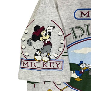 Puede incluir: Camiseta gris claro con Mickey Mouse jugando al golf. La palabra "MICKEY" est&aacute; en un cuadro rojo y azul. La camiseta tambi&eacute;n tiene las palabras "Mickey's" y "DI" en un dise&ntilde;o azul y verde. Tambi&eacute;n aparece el Pato Donald.