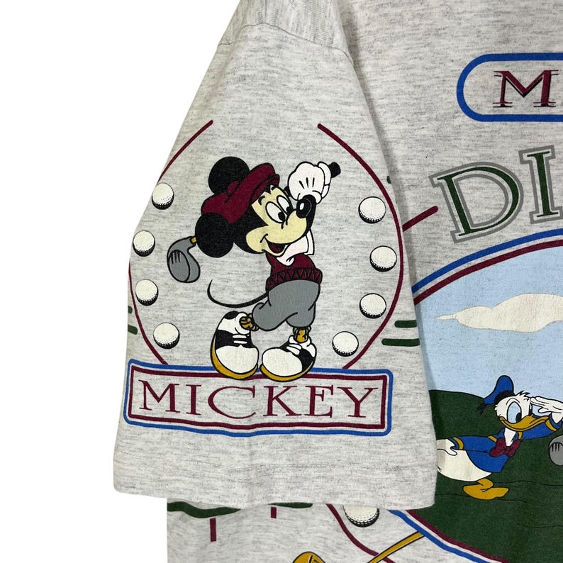 Puede incluir: Camiseta gris claro con Mickey Mouse jugando al golf. La palabra "MICKEY" est&aacute; en un cuadro rojo y azul. La camiseta tambi&eacute;n tiene las palabras "Mickey's" y "DI" en un dise&ntilde;o azul y verde. Tambi&eacute;n aparece el Pato Donald.