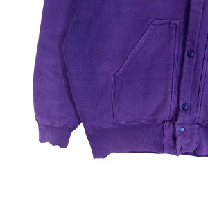 May include: A close-up of a purple sweatshirt with a button-up closure and a kangaroo pocket. The sweatshirt has a ribbed hem and cuffs. The buttons are a matching purple color.