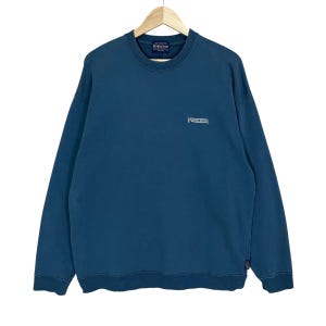 以下が含まれることがあります： 胸に白で「Pendleton」のブランド名が刺繍された、ティールブルーのクルーネックのスウェットシャツ。スウェットシャツは長袖で、裾と袖口はリブ編みです。木製のハンガーに吊るされています。