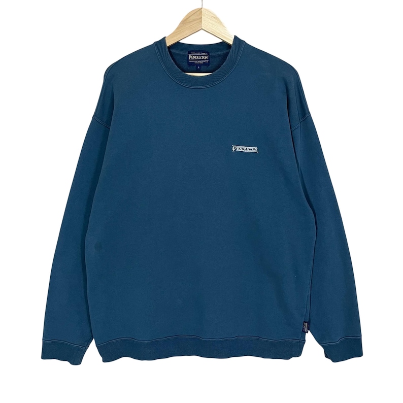 以下が含まれることがあります： 胸に白で「Pendleton」のブランド名が刺繍された、ティールブルーのクルーネックのスウェットシャツ。スウェットシャツは長袖で、裾と袖口はリブ編みです。木製のハンガーに吊るされています。