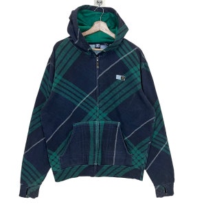 May include: A navy blue zip-up hoodie with a green and white plaid pattern. The hoodie has a green interior lining, a front pocket, and a small logo patch. The cuffs have thumb holes.