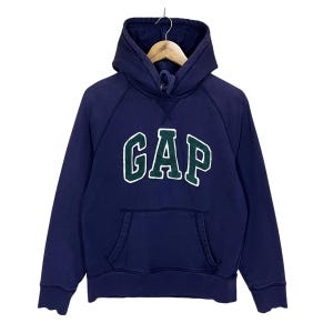 Puede incluir: Sudadera con capucha azul marino con la palabra "GAP" en letras grandes blancas y verdes en el pecho. La sudadera tiene un bolsillo de canguro y una capucha con cordón. La prenda está colgada de una percha de madera.