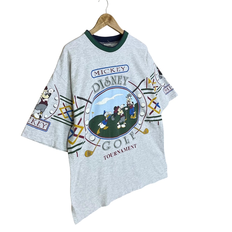 Puede incluir: Camiseta gris claro con un dise&ntilde;o de Mickey Mouse Disney Golf Tournament. La camiseta tiene cuello y ribetes de manga verdes. El gr&aacute;fico incluye a Mickey, Donald y Goofy jugando al golf en un campo verde con un fondo de cielo azul.