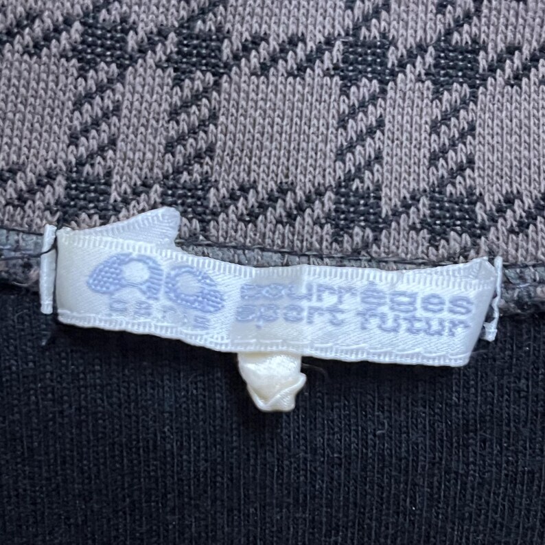 May include: Close-up of a garment tag with the text "Courr&egrave;ges Sport Futur" in light blue on a white label. The tag is attached to a black fabric, with a patterned gray and brown knit fabric above.