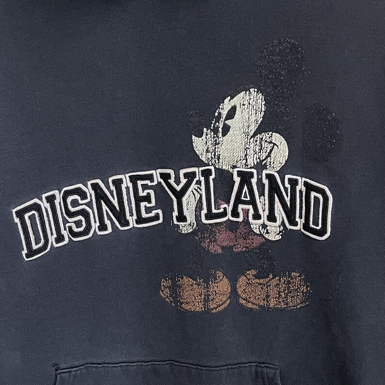 Puede incluir: Una sudadera gris oscura con un gr&aacute;fico de Mickey Mouse de estilo vintage y el texto "DISNEYLAND" impreso en la parte delantera.