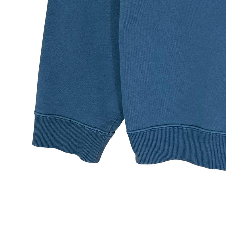 以下が含まれることがあります： ティールブルーのスウェットシャツのクローズアップ。画像は、裾と袖口を示しています。生地は柔らかいニット素材のようです。袖口と裾はリブ編みです。