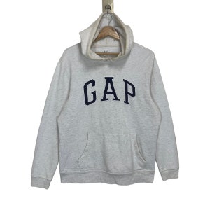 Puede incluir: Sudadera con capucha GAP gris claro con el logotipo arqueado azul marino en el pecho. La sudadera tiene una capucha con cordón, un bolsillo de canguro y mangas largas. La prenda está hecha de un material suave y cómodo.