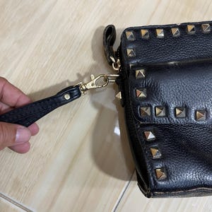 May include: Black leather clutch with gold-colored pyramid studs. The clutch has a wrist strap and a gold-colored clasp. The leather appears textured. The clutch is a small, rectangular shape.