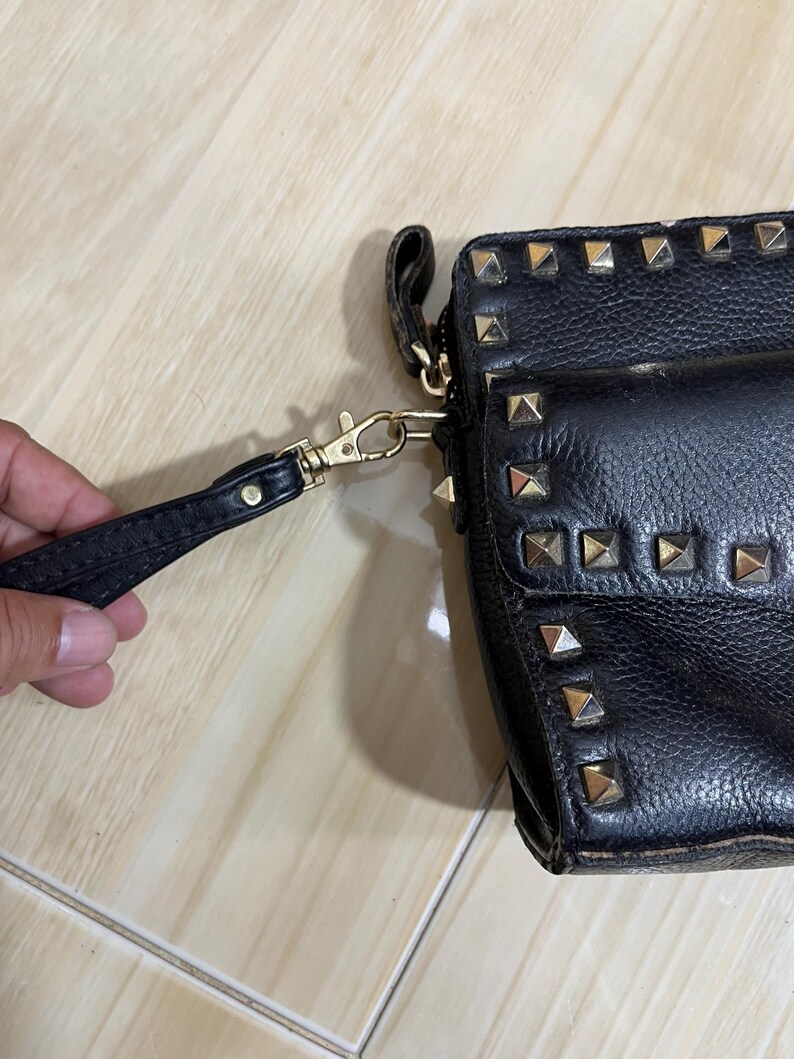 May include: Black leather clutch with gold-colored pyramid studs. The clutch has a wrist strap and a gold-colored clasp. The leather appears textured. The clutch is a small, rectangular shape.
