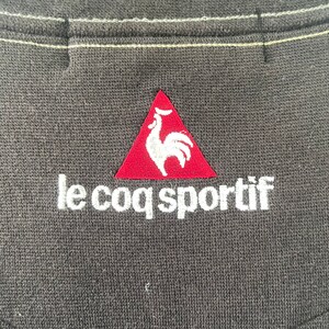 May include: A red triangle with a white rooster inside, and the text "le coq sportif" in white below the triangle. The logo is embroidered on a dark gray fabric.