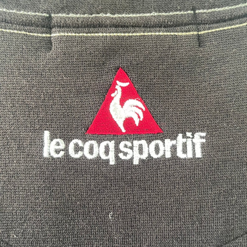 May include: A red triangle with a white rooster inside, and the text "le coq sportif" in white below the triangle. The logo is embroidered on a dark gray fabric.