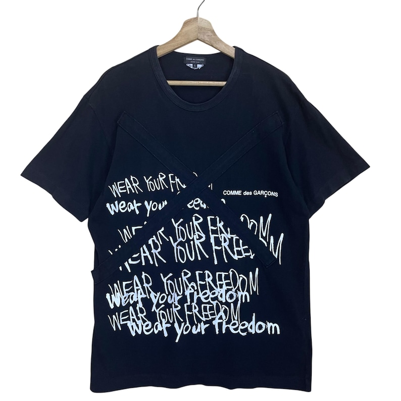 May include: Black short-sleeved t-shirt with white text that reads "Wear your freedom" repeated in different styles and sizes. The text is layered and appears to be written in a graffiti style. The shirt has a Comme des Gar&ccedil;ons label.