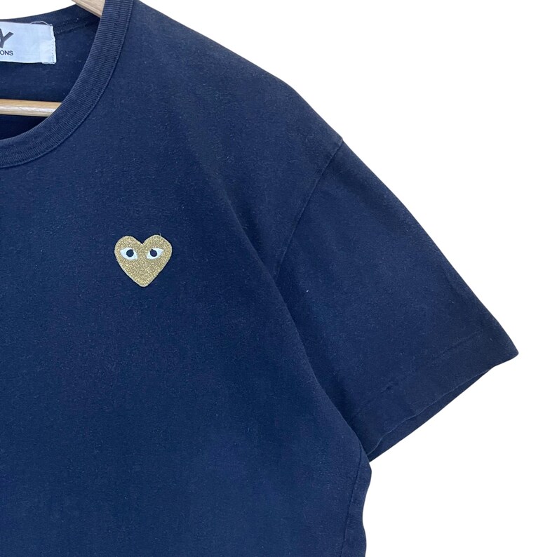 May include: Navy blue t-shirt with a gold heart-shaped emblem featuring two eye-like shapes. The shirt has a crew neck and short sleeves. The emblem is on the left chest area.
