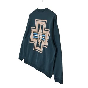 以下が含まれることがあります： 背面に幾何学的な十字のデザインが施されたティール色のスウェットシャツ。十字は、青、白、茶色の模様のベージュの縁取りが特徴です。スウェットシャツは柔らかい素材でできており、長袖です。