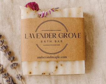 May include: A rectangular bath bar with a brown paper label that reads "LAVENDER GROVE BATH BAR" and "amberandmaple.com". The bar is decorated with dried flowers and lavender sprigs. The soap is off-white with visible flecks.