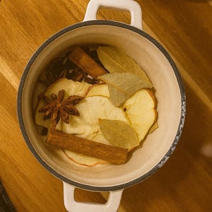 May include: A small, off-white ceramic bowl with two handles, filled with dried apple slices, cinnamon sticks, star anise, and bay leaves. The bowl sits on a wooden surface, with a white dish in the background.
