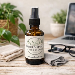 May include: A brown glass bottle of Amber & Maple screen cleaner with a black spray nozzle. The label features the brand name and a botanical design. A gray microfiber cloth and a laptop are in the background.
