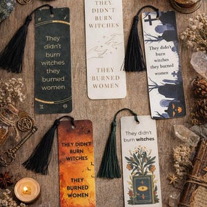 May include: A collection of decorative bookmarks with black tassels, featuring the text "They didn't burn witches they burned women." The bookmarks have various designs, including illustrations of hands, a graveyard, and a burning sunset. Candles and crystals surround the bookmarks.