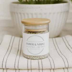 Könnte beinhalten: Ein klares Glas mit einem Holzdeckel, gefüllt mit Koriander-Limettensalz. Das Glas hat ein weißes Etikett mit dem Text "AMBER & MAPLE" und "CILANTRO LIME SALT". Das Glas steht auf einem gefalteten weiß-grau gestreiften Tuch.