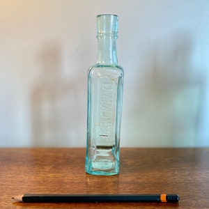 K&ouml;nnte beinhalten: Eine klare Glasflasche mit quadratischem K&ouml;rper und abgerundetem Hals. Die Flasche tr&auml;gt ein Etikett mit der Aufschrift "DR. BULL'S".