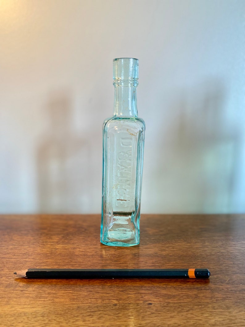 K&ouml;nnte beinhalten: Eine klare Glasflasche mit quadratischem K&ouml;rper und abgerundetem Hals. Die Flasche tr&auml;gt ein Etikett mit der Aufschrift "DR. BULL'S".