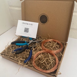 May include: A cardboard box filled with brown packing paper, two spools of copper wire, and a pair of blue and black pliers. The box has a sticker with the text "Wood & Wire Works" and a QR code on a card that says "Thank You!"