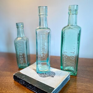 K&ouml;nnte beinhalten: Drei antike Glasflaschen mit einem Buch im Vordergrund. Die Flaschen sind klar und haben eine gr&uuml;ne T&ouml;nung. Die Flaschen sind mit "TON & GLY", "SAUCE" und "J. PETERSDORF" beschriftet.