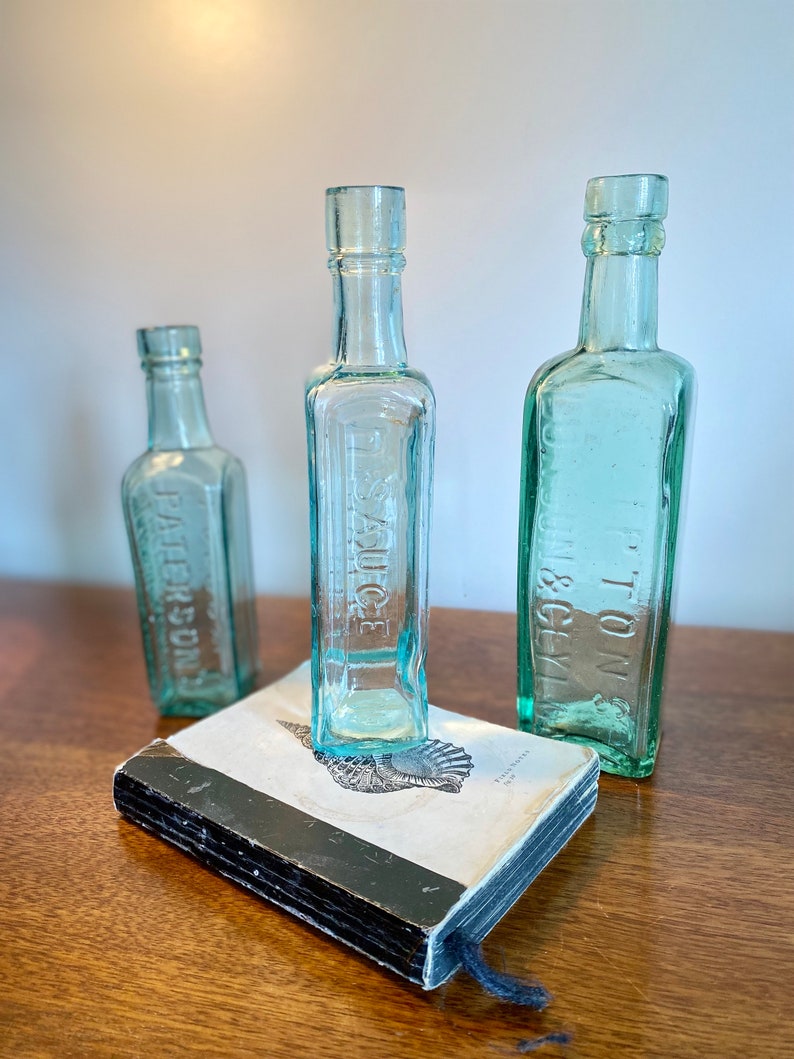 K&ouml;nnte beinhalten: Drei antike Glasflaschen mit einem Buch im Vordergrund. Die Flaschen sind klar und haben eine gr&uuml;ne T&ouml;nung. Die Flaschen sind mit "TON & GLY", "SAUCE" und "J. PETERSDORF" beschriftet.
