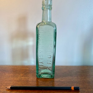 K&ouml;nnte beinhalten: Eine durchsichtige gr&uuml;ne Glasflasche mit quadratischem K&ouml;rper und schmalem Hals. Die Flasche tr&auml;gt den Text "S.S. TOWN & CITY" in Pr&auml;gung auf der Vorderseite.