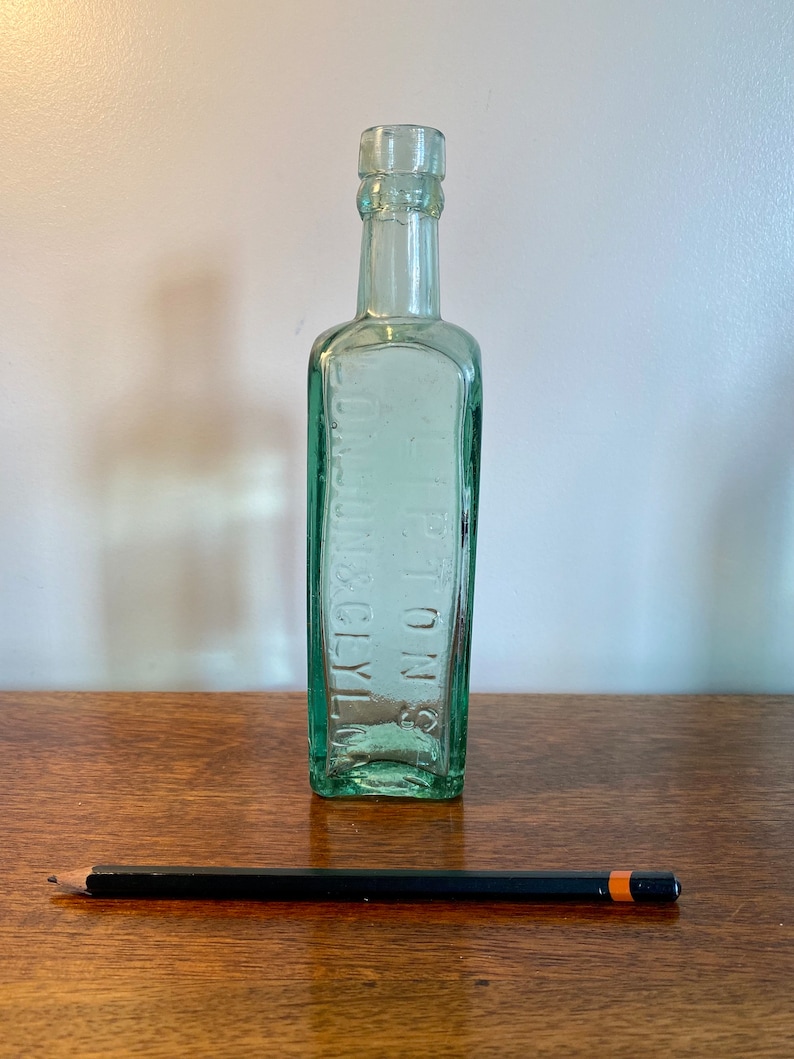 K&ouml;nnte beinhalten: Eine durchsichtige gr&uuml;ne Glasflasche mit quadratischem K&ouml;rper und schmalem Hals. Die Flasche tr&auml;gt den Text "S.S. TOWN & CITY" in Pr&auml;gung auf der Vorderseite.