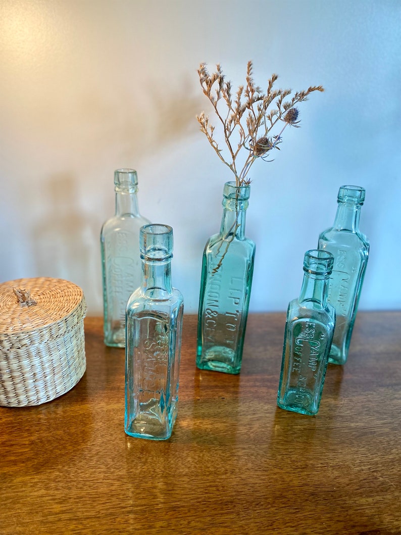 K&ouml;nnte beinhalten: F&uuml;nf klare Glasflaschen mit gepr&auml;gten Schriftz&uuml;gen auf einer Holzoberfl&auml;che. Die Flaschen sind mit getrockneten Blumen gef&uuml;llt und in einer Reihe angeordnet. Die Flaschen sind alle in verschiedenen Gr&ouml;&szlig;en und Formen.