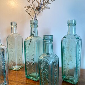K&ouml;nnte beinhalten: Sechs antike Glasflaschen mit gepr&auml;gter Beschriftung. Die Flaschen sind aus klarem gr&uuml;nem Glas und haben eine quadratische Form. Die Flaschen sind auf einer Holzoberfl&auml;che angeordnet. Die Flaschen sind alle unterschiedlich gro&szlig; und haben unterschiedliche Beschriftungen. Die Beschriftungen auf den Flaschen beinhalten die Namen von Unternehmen und Produkten. Die Flaschen sind alle in gutem Zustand und haben ein Vintage-Aussehen.