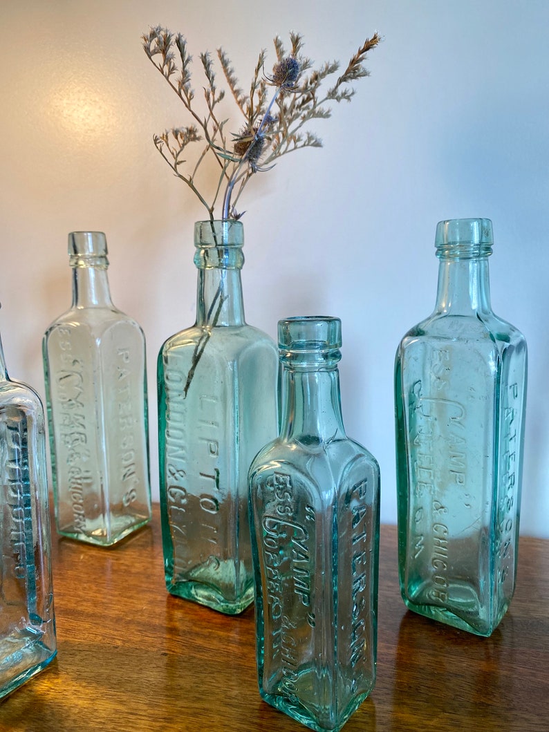 K&ouml;nnte beinhalten: Sechs antike Glasflaschen mit gepr&auml;gter Beschriftung. Die Flaschen sind aus klarem gr&uuml;nem Glas und haben eine quadratische Form. Die Flaschen sind auf einer Holzoberfl&auml;che angeordnet. Die Flaschen sind alle unterschiedlich gro&szlig; und haben unterschiedliche Beschriftungen. Die Beschriftungen auf den Flaschen beinhalten die Namen von Unternehmen und Produkten. Die Flaschen sind alle in gutem Zustand und haben ein Vintage-Aussehen.